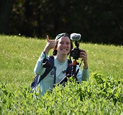 莉莉·克莱默一手拿着相机，一手竖起大拇指站在田野里