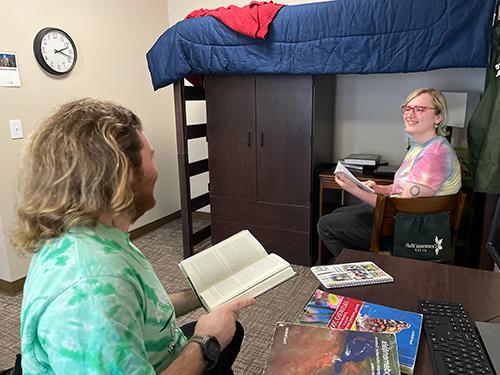 两名学生在百年三人间. 一个学生手里拿着一本书，另一个学生坐在双层床旁边的椅子上