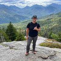 亚历克西斯·罗西奥·吉列尔莫身穿黑色t恤和灰色裤子站在岩石上. 背景是山脉